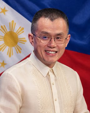 The image features a man wearing glasses, smiling at the camera with his hands placed behind his back. He is dressed in a white shirt that has lace detailing. The background of the photo includes a flag from the Philippines, which is blue, red, and yellow colors. There are also stars on the flag. The man's attire and the presence of the Philippine flag suggest that he might be attending an event or celebration related to his country.