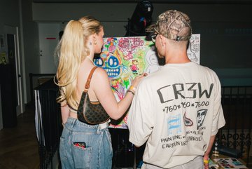 The image shows two people standing next to each other, with one person wearing a white shirt that reads "Crow W" and another person wearing a camo hat. They appear to be engaged in an activity related to art or painting. The background of the photo is blurred, but it seems like they are inside a room where there might be more artwork displayed on the walls. There's also a handbag visible near one of them. The image includes text that reads "Solana Summer House LA did something to people and these photos are proof spot a friend?". This suggests that the two individuals in the photo may have been involved in an art project or event, possibly at Solana Summer House.