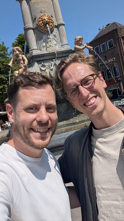 A selfie of two smiling men standing in front of an ornate fountain with cherub statues and a 'Chicago' sign visible in the background.