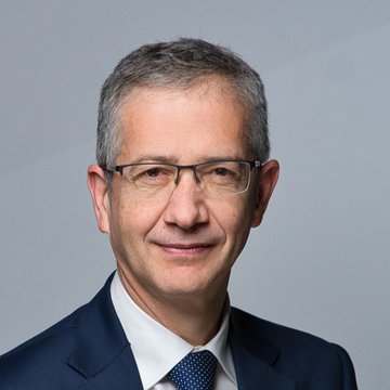 A professional headshot of a man with gray hair, wearing rimless glasses, a dark suit, white shirt, and dark patterned tie, smiling subtly against a gray background—identified as BIS chief Pablo Hernández de Cos, who recently argued that stablecoins lack credibility as a scalable payment method compared to tokenized bank deposits.