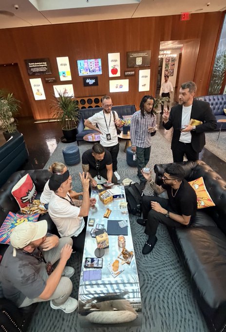 A group of men gather around a coffee table in a lounge area with framed awards on the wall behind them, some sitting on couches and others standing, appearing to socialize with drinks in hand.