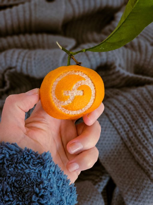 A hand holding a small orange with the Google 'G' logo carved into its peel, set against a gray knitted blanket background.