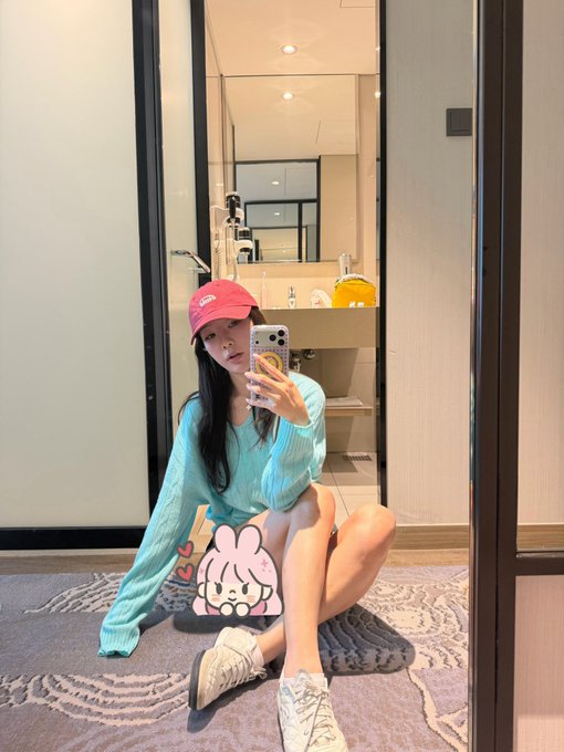 A young woman in a light blue sweater and red cap takes a mirror selfie while sitting on the floor of a modern bathroom, with a cute bunny sticker edited onto the image. The caption ties into the post's "weekend checklist" theme, evoking a sense of relaxed downtime and personal time off—though notably, opening the Binance app isn't actually depicted in the image.