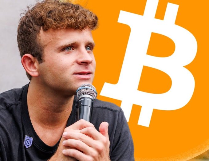 A man with curly hair speaking into a microphone in front of a large orange Bitcoin logo, appearing to be at a cryptocurrency-related event.