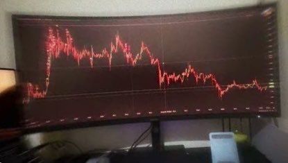 A curved computer monitor displays a red line stock chart on a black background, showing a sharp decline followed by volatility in the dark room.