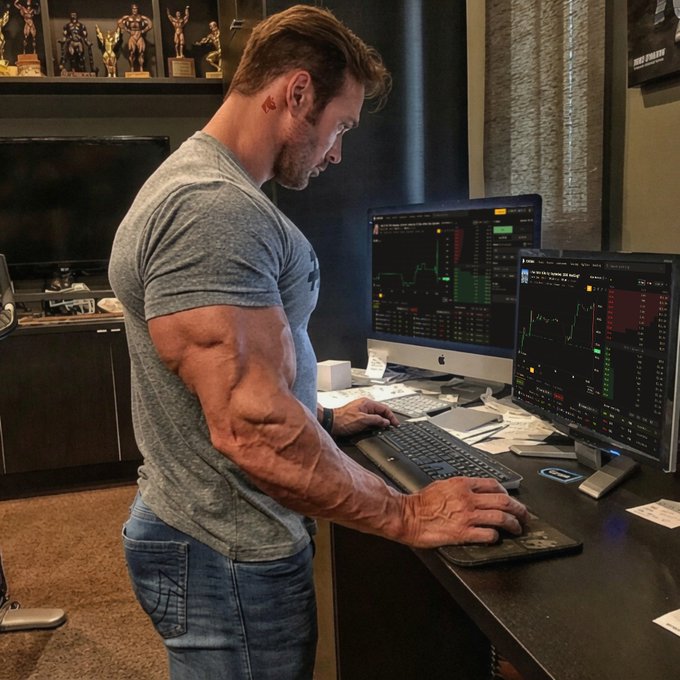A side profile of a muscular man in a gray t-shirt standing at a desk using a keyboard and mouse while analyzing financial charts on dual monitors, with bodybuilding trophies displayed on a shelf in the background.
