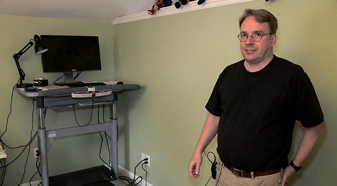 A man wearing glasses, a black short-sleeved shirt, and khaki pants stands in a room with green walls next to a standing desk holding a monitor and lamp.