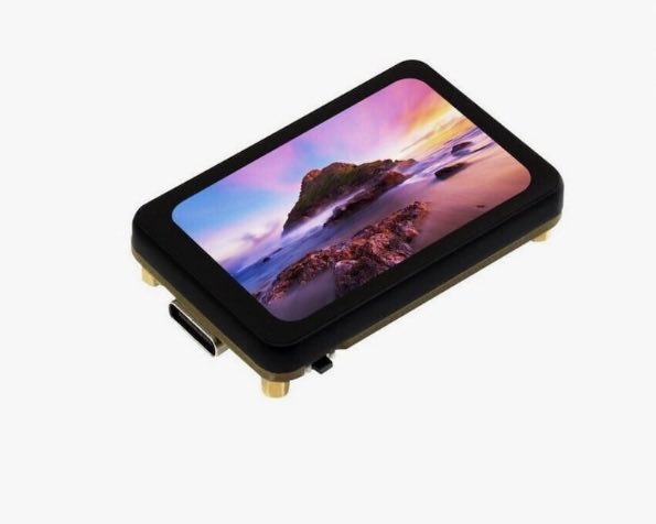 A small rectangular electronic display module with a black casing and gold-colored mounting pins, showing a scenic landscape wallpaper of a rocky coastline at sunset.
