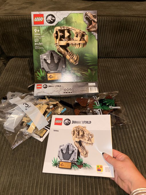 A LEGO Jurassic World 76964 dinosaur skull set is unboxed on a couch, with the box, instruction manual, and bags of tan and brown pieces spread out.