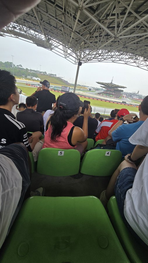 Spectators sit in a covered seating area with green chairs marked "5" and "4," watching a race at what appears to be the Sepang International Circuit in Malaysia, with the track's distinctive control tower visible in the distance. The image captures the rare moment of enjoying a complimentary F1 experience courtesy of a generous crypto friend, as referenced in the caption about receiving free tickets from a semi-retired acquaintance.