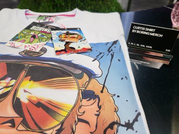 A close-up of clothing items including a graphic t-shirt with sunglasses artwork and stickers, next to a tag reading 'CURTIS SHIRT BY YOUNG MERICH'.
