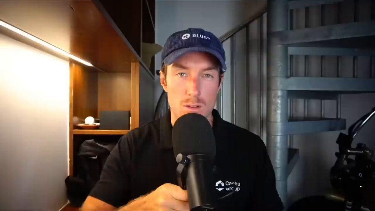 The image shows a man wearing a blue baseball cap holding a microphone, possibly recording or broadcasting something related to Ripple Coin. The background features a staircase, suggesting that he might be in an office or studio setting. The caption reads "People are waking up to XLM... wait until they hear what the BIS has been doing with Ripple".