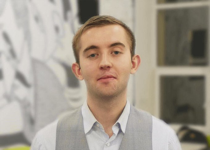 A young man with short brown hair wearing a light blue checkered shirt and gray vest, looking at the camera in an office setting.