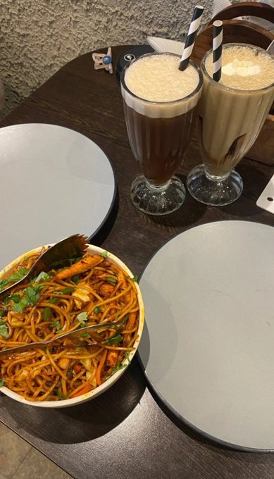 A table setting with two tall glasses of latte, a bowl of stir-fried noodles with vegetables and shrimp, and two empty plates ready to be served.