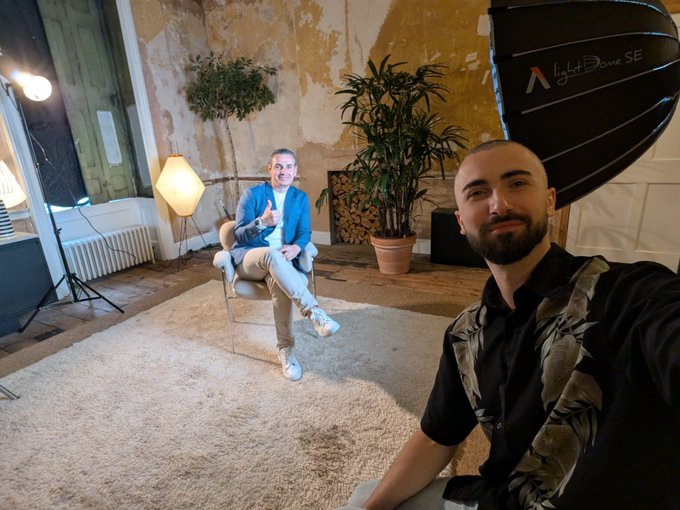 A selfie-style photo taken in a photography studio with an octagonal softbox (light Dome SE) visible, showing two men - one seated in a blue blazer and one in the foreground wearing a dark shirt with leaf patterns.