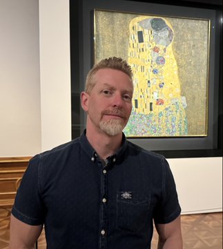 A man with short hair and a goatee wearing a dark blue button-up shirt poses in front of a framed Gustav Klimt painting reminiscent of 'The Kiss'.