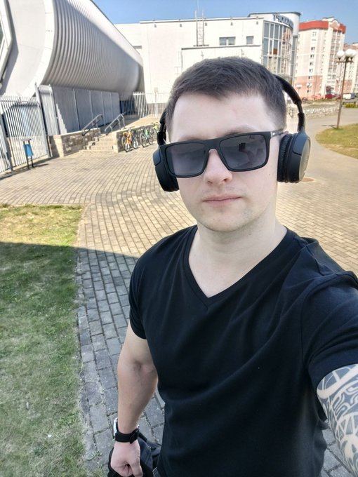 A selfie of a man wearing a black v-neck t-shirt, sunglasses, and over-ear headphones, standing outdoors on a paved walkway near a modern building.