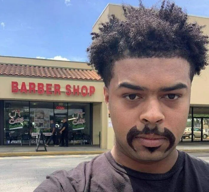 A young man takes a selfie in front of 'JT Cuts Barber Shop', displaying a freshly styled mustache with curled handlebars while looking disappointedly at the camera.