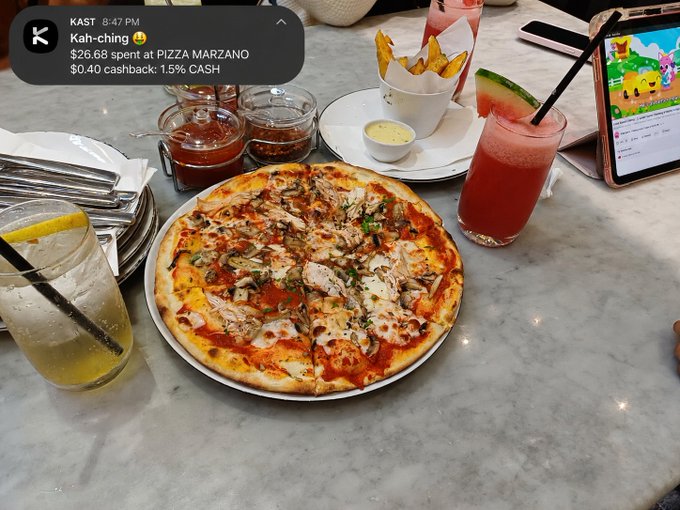 A meal at Pizza Marzano with a pizza, drinks, and condiments on a marble table, showing a notification of a $26.68 payment with $0.40 cashback.