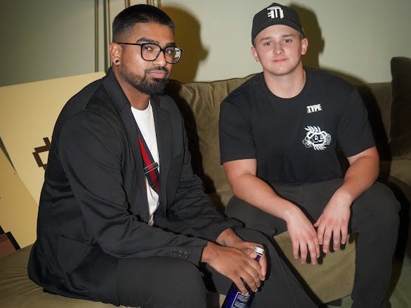 Two men sitting side-by-side on a couch; the man on the left wears glasses, a black jacket, and a plaid shirt, while the man on the right wears a black cap and t-shirt with logos.