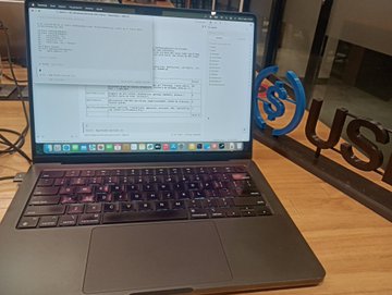 A MacBook laptop sits open on a wooden desk displaying lines of code on its screen, with a 'SUSU' sticker or logo visible on a glass partition in the background.