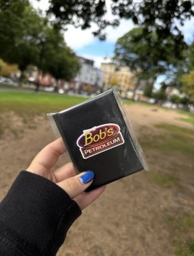 A hand with blue nail polish holds a black wallet featuring a 'Bob's PETERSFIELD' logo, displayed against an outdoor park background.