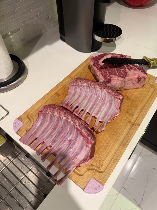 A rack of raw lamb on a wooden cutting board with a knife, with a larger cut of meat in the background, sits ready for preparation on a kitchen counter. The image visually represents the "86K reasons to feast" caption by showcasing a hearty, indulgent meal in the making—likely highlighting the value or satisfaction of a generous spread of food.