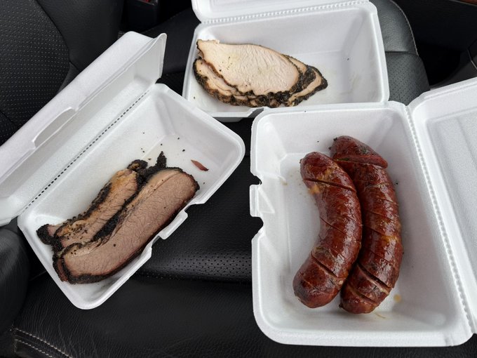 Takeout containers hold sliced smoked brisket and two sausages, showcasing a hearty carnivore lunch from Black Gold BBQ.