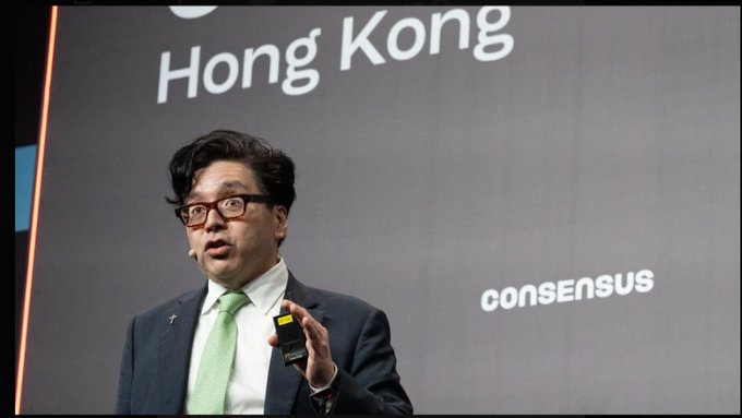 A man in a dark suit, green tie, and red glasses speaking on stage at a 'consensus' event in Hong Kong, holding a presentation clicker.