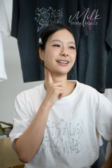 A smiling young woman wearing a white graphic t-shirt rests her chin on her hand indoors.