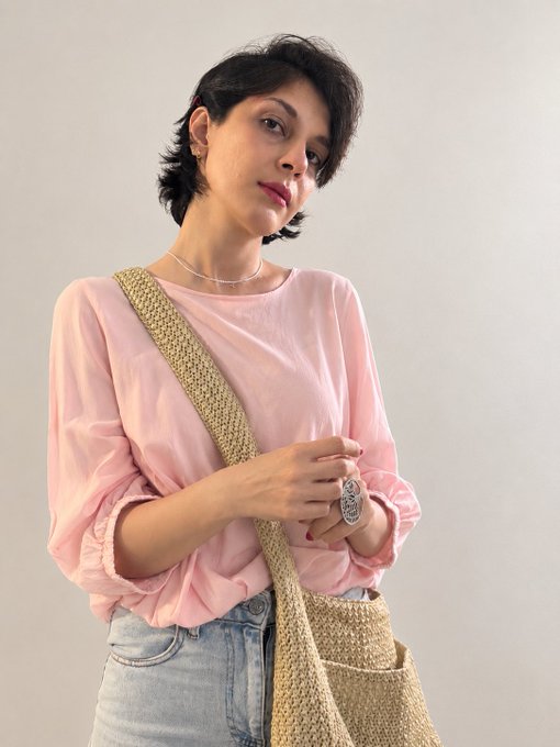 A woman with short dark hair poses wearing a loose pink top, light blue jeans, and a woven crossbody bag strap, accessorized with a silver necklace and a large ring.