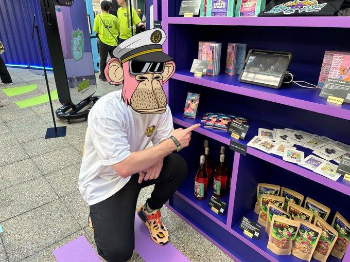 A person wearing a Bored Ape-style captain hat mask, sunglasses, and a white t-shirt poses at a Bored Ape-themed merchandise display featuring books, bottles, and collectibles on purple shelves.