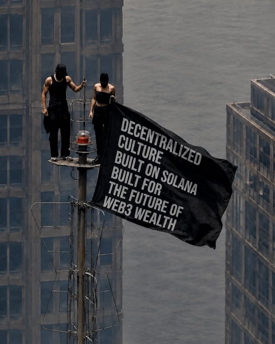 Two figures dressed in black stand atop a communications tower holding a large black banner reading 'DECENTRALIZED CULTURE BUILT ON SOLANA BUILT FOR THE FUTURE OF WEB3 WEALTH' against a city skyline.