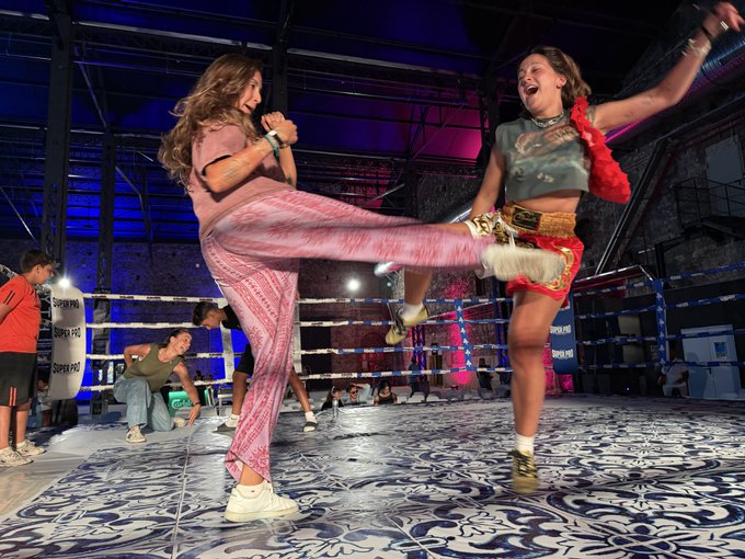 Two young women playfully fighting inside a boxing ring with patterned mat flooring; one in pink pants is mid-kick while the other, wearing a championship belt and red boa, laughs with arms raised.
