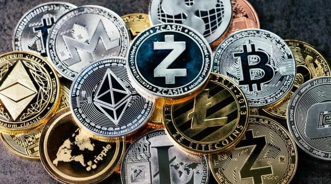 A pile of various physical cryptocurrency coins, including Bitcoin, Monero, Zcash, Ethereum, Ripple, and Litecoin, arranged on a dark surface.