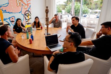 A group of professionals gathers around a wooden table in a modern meeting space, engaged in discussion with a laptop and drinks visible, while colorful abstract artwork decorates the wall behind them. The setting reflects the post's theme of collaborative dialogue with EEA community leaders, capturing the informal yet purposeful exchange of feedback to shape product development.