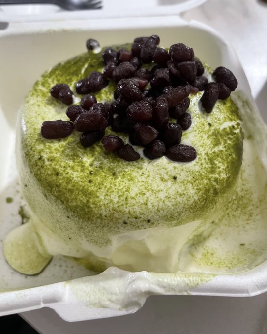 The image features a small, round pastry with a white base and green frosting topped with black beans. The pastry appears to be covered in sauce or dressing as well. The background of the image is blurred, drawing attention to the main subject - the delicious dessert.