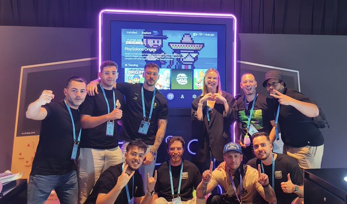 A group photo of eight people posing enthusiastically in front of an 'ORIGIN' digital display screen, with some making hand gestures like heart shapes and shaka signs.