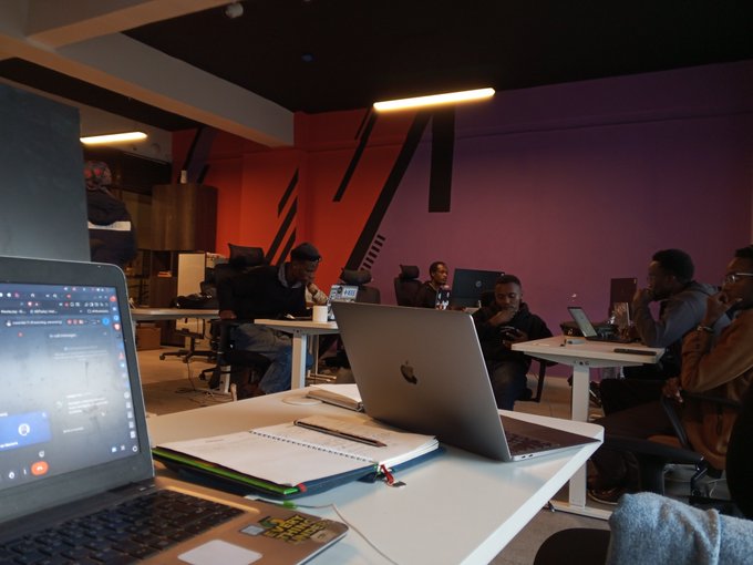 Collaborators gather around laptops during a meeting in a modern office space with pink and red walls, working together on community growth strategies and KPIs.
