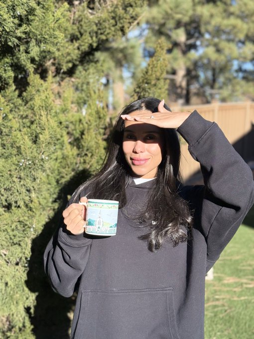 A woman in a black hoodie holds a mug while shielding her eyes from the sun outdoors near some trees.