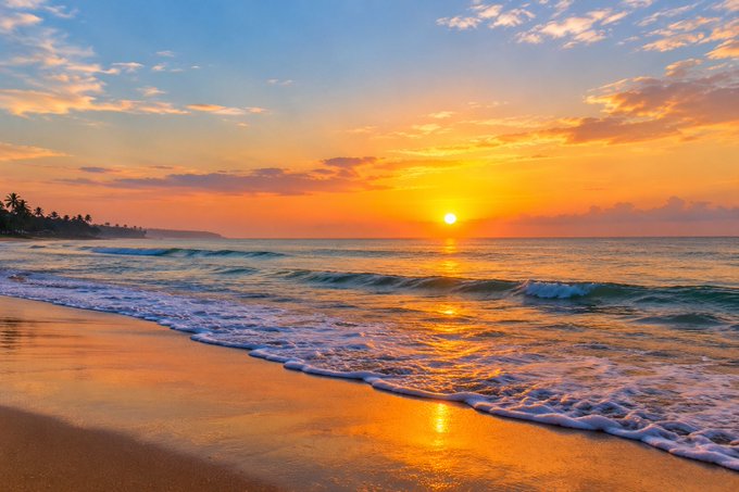 A vibrant sunset over the ocean with golden and orange hues reflecting on the waves washing onto a sandy beach.
