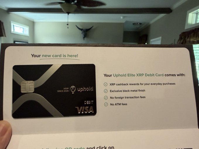 A person holds up a Uphold Elite XRP Debit Card welcome package, showcasing its black metal finish and listing benefits including XRP cashback rewards, no foreign transaction fees, and no ATM fees. The caption's excitement at receiving the @UpholdInc card aligns with the "Your new card is here!" message prominently displayed on the holder.