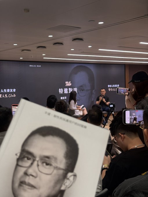 A person holds a book featuring a black-and-white photo of a man's face in the foreground, while in the background a panel discussion takes place on a stage with the same man's image projected on a large screen.
