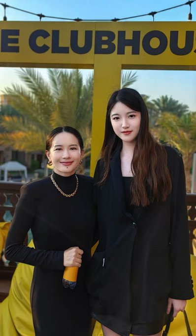The image features two women standing next to each other, both dressed in black clothing. They are posing for a photo together, with one woman holding an object that appears to be a bottle or a cup. The background of the image includes a yellow sign, which is likely related to the event they are attending.
