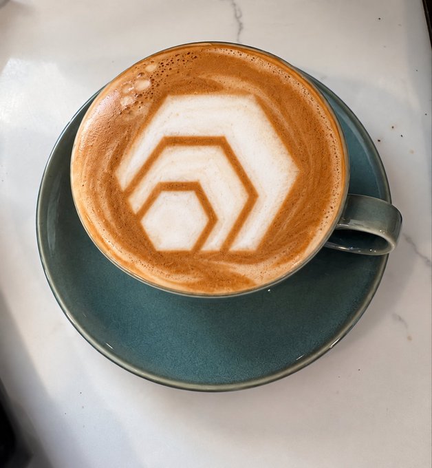 A cappuccino in a teal ceramic cup features intricate latte art of layered hexagons, served on a matching saucer on a marble surface.