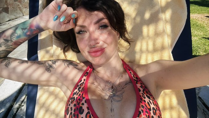A woman with freckles, multiple tattoos on her arms and chest, and blue nail polish poses in a leopard-print bikini while lounging outdoors.