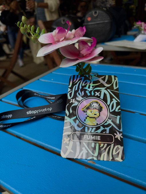 A laminated badge with a pixel-art character and the name "FUMIE" rests on a blue table beside pink freesia flowers and a Doginals Dogs lanyard, marking the meetup.