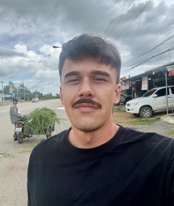 A young man with a mustache taking a selfie on a roadside, with a person on a motorcycle loaded with green grass in the background and parked white pickup trucks nearby.