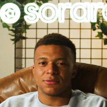 The image features a man sitting in a chair, wearing a white t-shirt. The background of the photo shows a neon sign that says "Sorair" written in white letters. There are also some plants visible behind the man. The main subject is the man who appears to be posing for the picture.