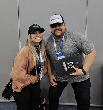 Two people pose together at an event, with the woman wearing a black trucker hat featuring the "ARO" logo and a lanyard, while the man wears a white "ChainCatcher" cap and holds a card also displaying the ARO logo—visually aligning with the post about Paula representing the ARO Network at Blockchain Rio.